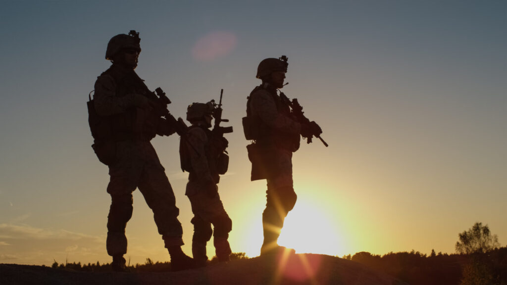 Squad of Three Fully Equipped and Armed Soldiers Standing on Hill in Desert Environment in Sunset Light.