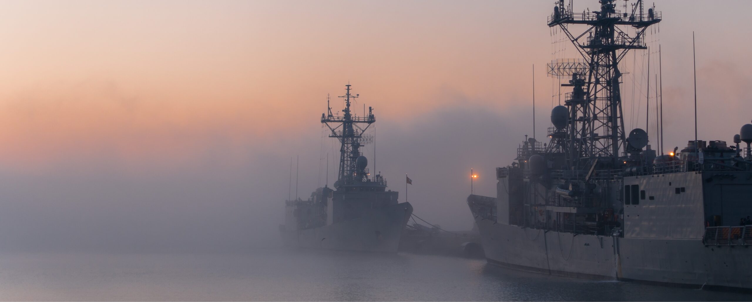 Two naval ships docked near a foggy harbor at sunset with a calm sea and a gradient sky ready to deploy mission military drones.