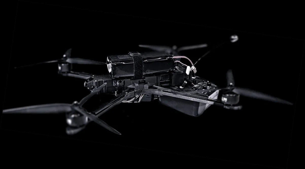 Black quadcopter military drone with four propellers and visible wiring, shown against a dark background.